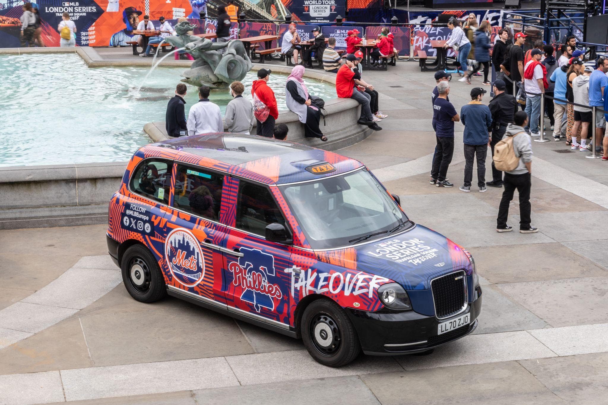 A Major League Baseball wrapped black cab beside the Trafalgar Square fountain, surrounded by event crowds